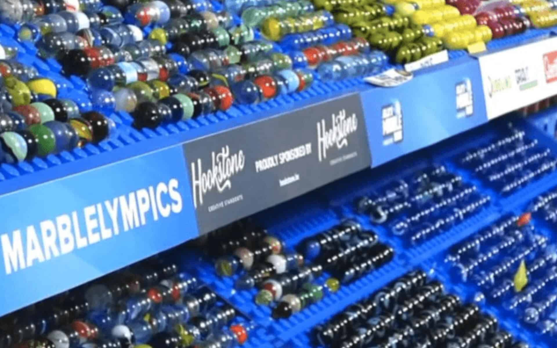 A marble display shows various shooters and alleys in a blue plastic rack with a sign reading "marblelympics"